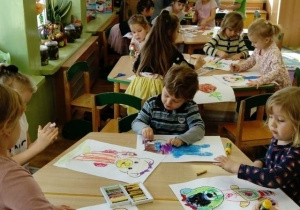 Dzieci siedzą przy stolikach i kolorują kontur misia.